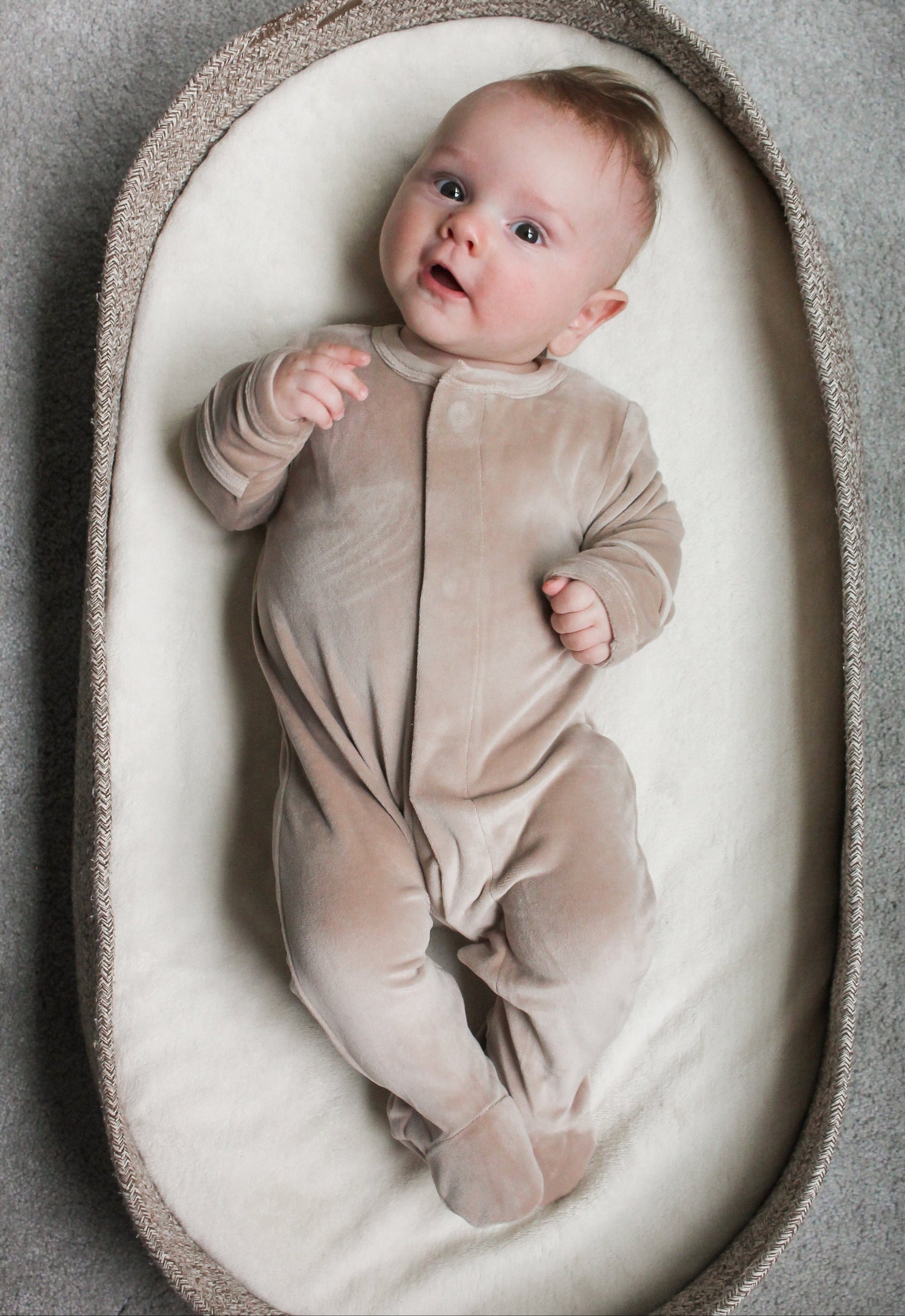 Baby in a beige onesie lying in a textured baby nest on a gray background